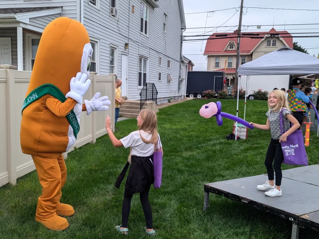 A mascot whose costume is shaped like a Long John donut is speaking to a child. Off to the right, another child stands on a temporary stage with a balloon shaped into a flower. Both children hold purple tote bags.