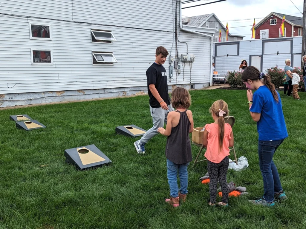 Three children face a set of four cornhole boards in the grass of the New Holland town centre. A teen walks among the boards.