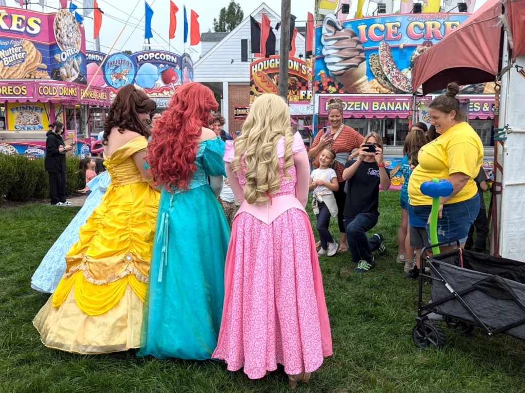 Three women face away from the camera: one wearing a bright yellow dress, one in a a teal blue dress, and one with a bubblegum pink dress. They appear to be posing for a photo with people in the background aiming cameras at them.