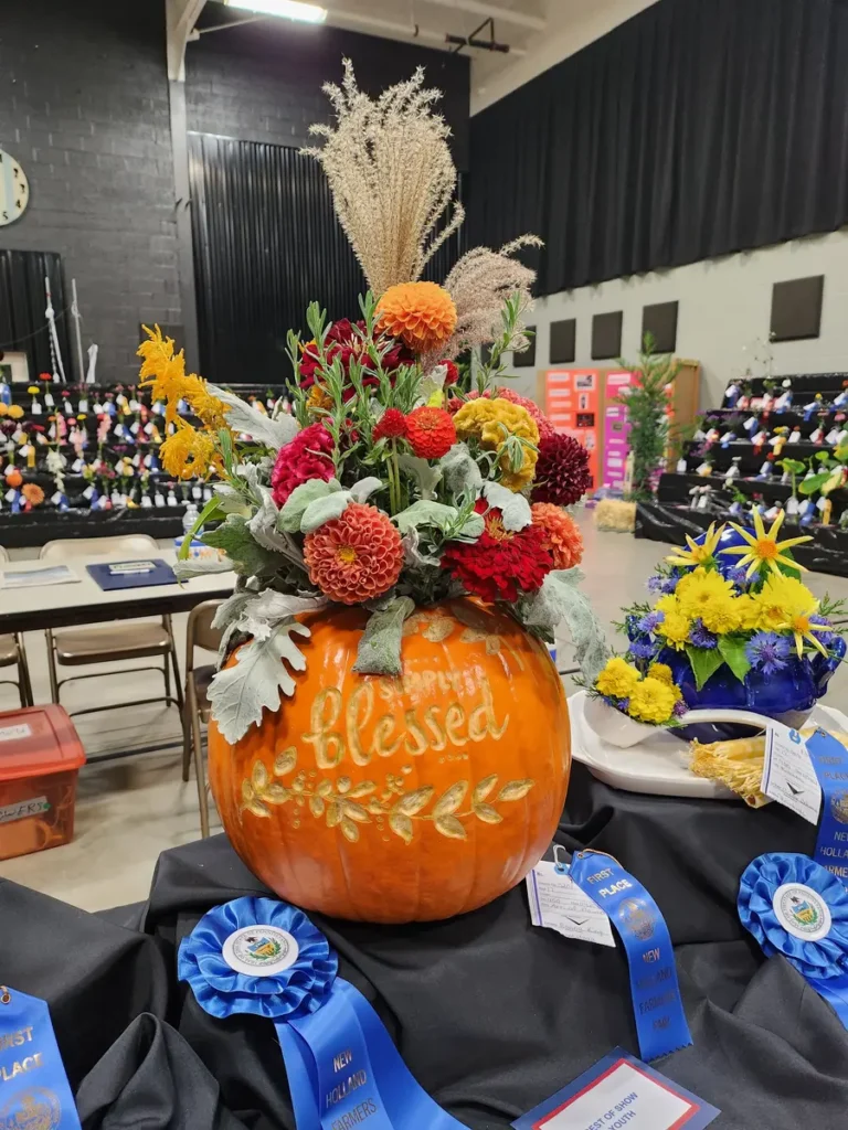 Flowers arranged in a pumpkin with “blessed” decoratively carved into it and a best-in-show rosette laying near it.