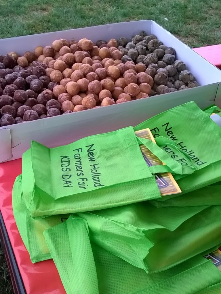 Hundreds of donut holes in a box next to a stack of green reusable bags labeled "New Holland Farmers Fair Kids Day"