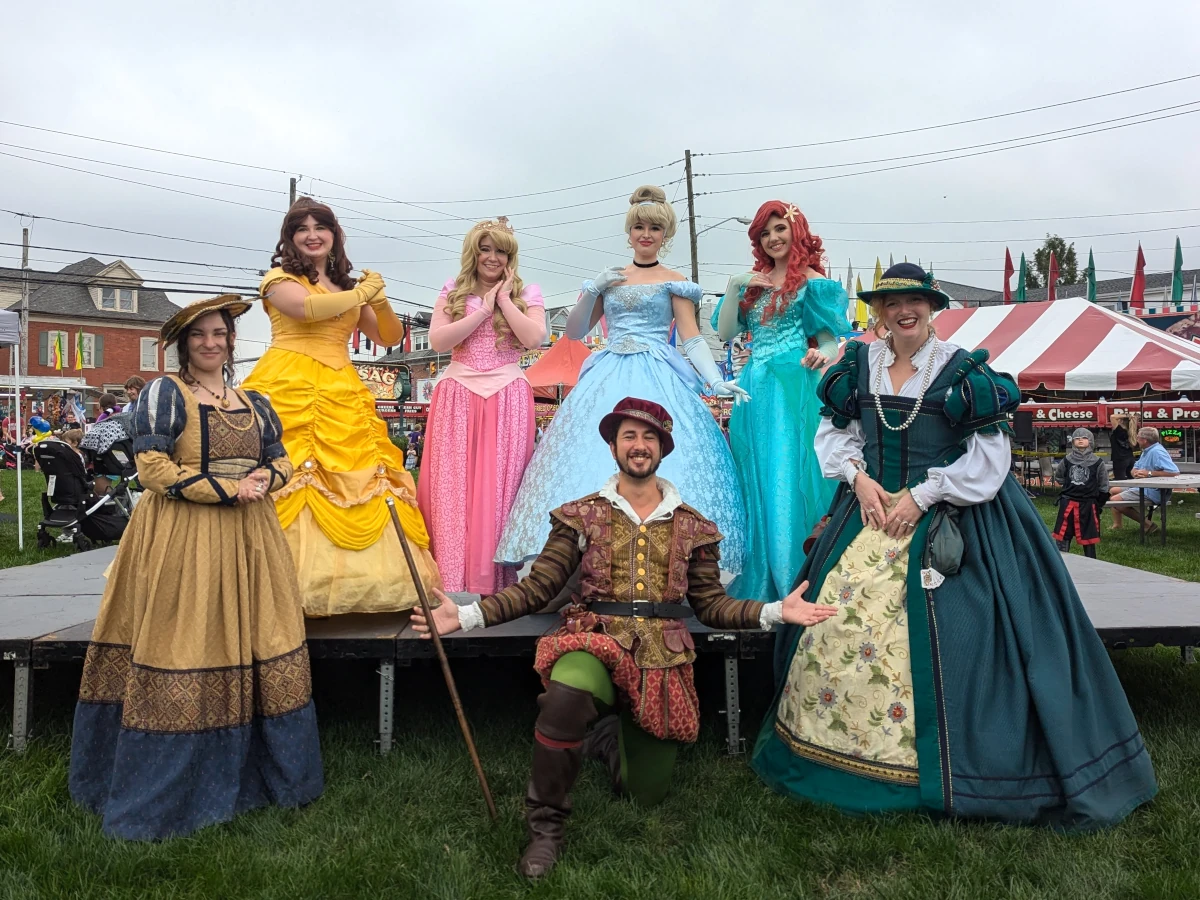 Seven people pose for a photo.Four of them are women standing in back on a raised stage with brightly colored dresses: the leftmost wears a yellow dress, then bubblegum pink with a tiara, then blue, and finally a teal dress with a starfish hair clip. In front of them are the other three: a man kneeling in a ren faire outfit with walking stick flanked by two standing women in ren faire dresses.