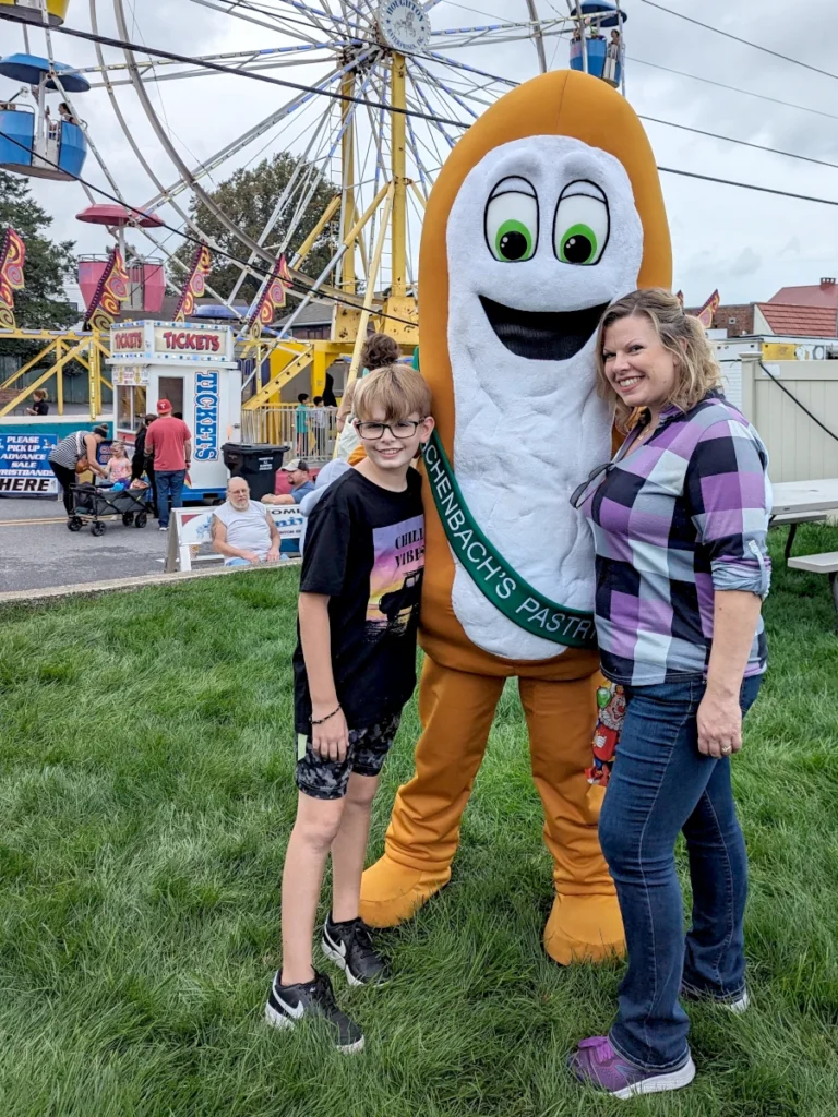 A mascot shaped like a long john donut wearing a sash that reads "Auchenbach's Pastries" poses with its arms around a child and an adult. In the background is a ferris wheel.