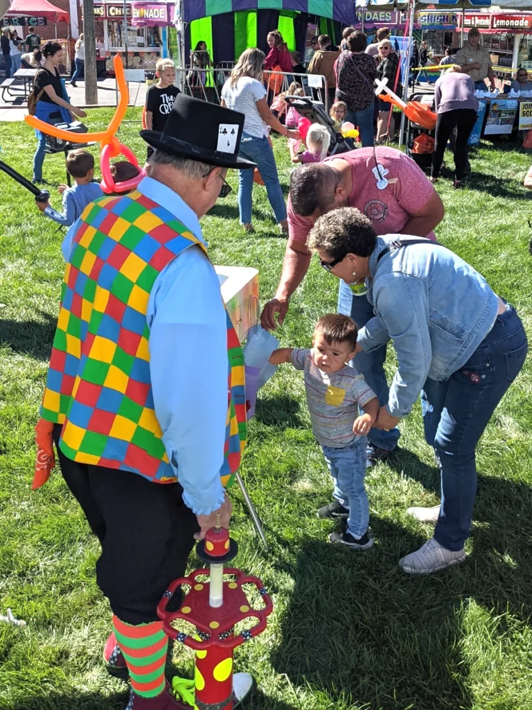 A clown facing away from the camera wearing a bright, colorful vest of green, blue, red, and yellow checkers and a top hat with a playing card on the brim. A child is reaching into a bag with the assistance of two adults. In the background, children play with shaped balloons. A balloon pump sits on the ground by the clown's feet.