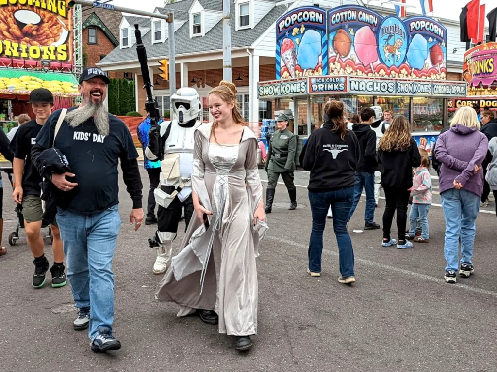 A woman in a dress is accompanied by a storm trooper and others in military uniforms walking through the fair midway with concession stands in the background. A man walks near the woman wearing a black T-shirt labeled "KIds' Day."