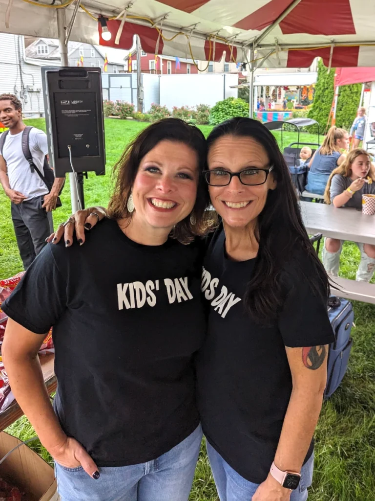Two women stand, one with her arm around the other. They wear matching black T-shirts that read "Kids' Day."